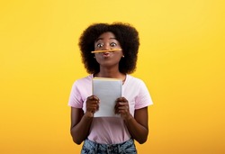 Funny black female student keeping pencil between nose and lips as moustach and holding notebook, posing over yellow studio background, copy space