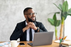 Portrait Of Pensive African Business Man In Glasses Sitting At Desk With Laptop In Home Office, Looking Away With Glad Face Expression. Smiling Guy In Suit Thinking About Successful Business Strategy