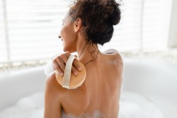 Back view of young woman making lymphatic massage with brush, scrubbing her skin in foamy bath at home. Millennial lady brushing her back, taking care of body in bathtub, indoors