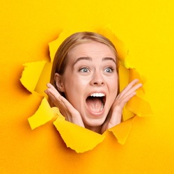 Beautiful blonde woman shouting OMG, shocked to learn about huge sale or discount, looking through hole in ripped orange paper. Amazing offer, great advertisement concept