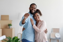 Happy smiling young african american loving family hugging in new home among boxes and showing house keys, man and woman spouses feel joy. Moving, relocating to own apartment, rental and real estate