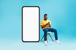 Cheerful young black man sitting on chair near big smartphone with empty screen, using mobile device, presenting space for website, advertisement or mobile app, blue studio background