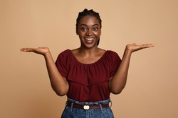 Choice, choosing, balance concept. Confused african american young woman holding something invisible on both palms, trying to choose something, holding hands up as scales, beige studio background