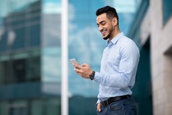 Positive middle eastern guy in formal outfit standing by modern business center, using smartphone and smiling, sending emails while having break, panorama with copy space, side view