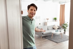 Welcome. Portrait of cheerful man inviting visitor to enter his home, happy young guy standing in doorway of modern apartment, millennial male holding door looking out showing living room with hand