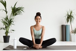 Smiling Beautiful Woman Sitting On Yoga Mat And Listening Music On Smartphone With Earphones, Happy Brunette Female Stretching Or Practicing Meditation And Enjoying Her Favorite Playlist, Copy Space