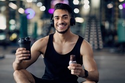 Fitness Nutrition. Happy Muscular Arab Man Holding Sport Shaker And Protein Bar While Relaxing After Training At Gym, Smiling Middle Eastern Male Athlete Having Break In Workout, Enjoying Snacks