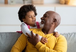 Cheerful little black girl play with older grandfather from back, hugging man at home interior. Family, people, relationships of generations during covid-19 lockdown. Positive active at living room