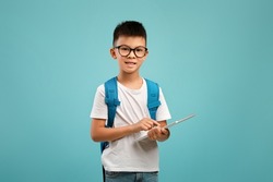 Gadgets For Study. Cute Asian Schoolboy In Eyeglasses Holding Digital Tablet And Looking At Camera, Korean Preteen Boy Wearing Backpack Using Tab Computer For Online Learning, Blue Background