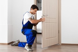 Installation of a lock on the front wooden entrance door. Portrait of young locksmith workman in blue uniform installing door knob. Professional repair service. Maintenance Concept