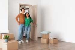 Relocation Concept. Happy millennial newlyweds walking in new empty house with cardboard boxes on floor, young family of two people moving into bought apartment, excited guy and lady choosing flat
