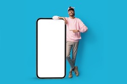 Stylish indian guy standing by huge smartphone with blank screen, mockup, pointing at advertisement, smiling millennial man recommending newest mobile application, blue studio background, copy space