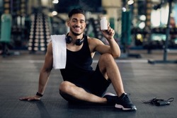 Young Muscular Arab Man Holding Container With Supplement Pills, Middle Eastern Male Athlete Posing At Gym With Blank Pack Of Multivitamins In Hands, Sitting On Floor In Sport Club And Smiling