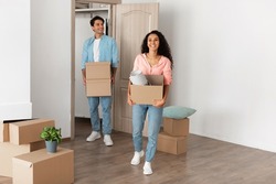 Relocation Concept. Happy millennial newlyweds holding cardboard boxes walking in new house, young family of two people moving into bought apartment, excited guy and lady carry personal belongings