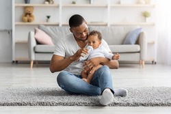 Childcare Concept. Loving African American Father Giving Bottle With Water To Baby Son At Home, Black Young Dad Spending Time With His Cute Infant Child, Sitting On Floor In Living Room, Copy Space