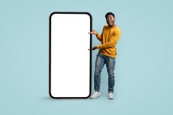 Cheerful african american millennial man showing huge modern cellphone with blank screen, showing newest entertaining or dating mobile app, mockup, blue studio background, copy space