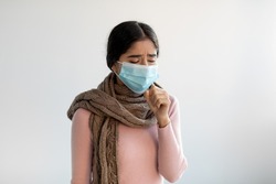 Upset despaired young indian lady in protective mask and scarf coughs, feeling bad and suffers from illness on gray background. Flu, cold, covid-19 virus and health problems. New normal, quarantine