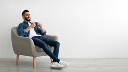 Full length of young Arab man drinking hot coffee in armchair against white studio wall, banner with free space. Peaceful middle Eastern guy having relaxing day, chilling on lazy morning