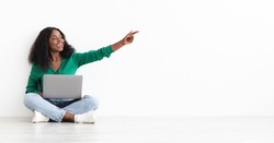 Emotional young black woman in casual sitting on floor by white wall with modern laptop, showing empty space, happy pretty african american lady surfing on Internet, shopping online, panorama