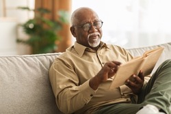 Senior African American Man Reading Book Sitting On Couch At Home, Wearing Eyeglasses. Retired Male Enjoying Reading New Novel Or Business Literature On Weekend. Retirement Leisure Concept