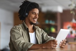 Happy Black Guy Wearing Wireless Headphones Using Digital Tablet At Cafe, Smiling Young African American Man Relaxing With Tab Computer While Sitting At Table In Modern Coffeeshop, Copy Space
