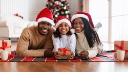 Merry Christmas And Happy New Year. Portrait Of Happy Loving Black Family In Santa Claus Hats Celebrating Winter Holiday Together, Lying On Floor Blanket In Decorated Living Room, Posing At Camera