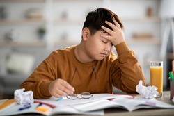 Exhautsed chubby asian kid teen boy doing homework alone, sitting at table full of exercise books, notepads, glass of orange juice, touching his head, experiencing difficulties with studying