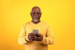 Happy senior black man using smartphone, texting to family, sending sms, browsing web on orange studio background. Elderly African American male checking social network on mobile device