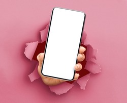 Mobile App, Great Offer. Closeup of female hand holding smartphone with white empty screen showing device close to camera breaking through pink paper sheet. Gadget display with free copy space, mockup