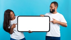Mobile App Advertisement. Excited Black Couple Holding And Pointing At Big White Empty Smartphone Screen, People Presenting Cell Phone Display Mock Up On Blue Studio Background. Check This, Panorama