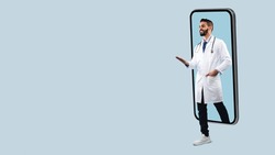 Telemedicine. Young Happy Male Middle Eastern Doctor In White Uniform With Stethoscope Coming Out Big Cell Phone Screen, Stepping Walking Off Gadget Monitor, Showing Free Copy Space, Panorama Banner