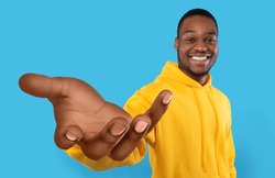 Happy African American guy showing big outstretched hand, offering help, taking or giving something, reaching out for support on blue studio background. Selective focus
