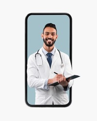 Medical Services. Young Happy Male Middle Eastern Doctor In White Uniform With Stethoscope And Clipboard Writing Prescription To Patient And Smiling At Camera, Looking Coming Out Big Cell Phone Screen