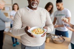 African american senior man holding plate with meal, receiving food in charity organization office, cropped, closeup. Volunteers helping feed elderly and homeless people with free meals