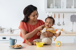 Childcare. Loving Black Mom Feeding Her Cute Baby Son From Spoon In Kitchen, Caring Mother Giving Porridge Or Mash Fruit Puree To Adorable Infant Child At Home, Preparing Healhy Food For Kid