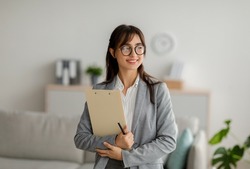 Portrait of female arab psychotherapist with clipboard looking aside and smiling, standing at modern office, free space. Professional psychoanalyst providing psychological services at clinic