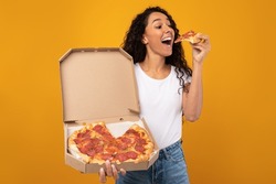 Excited Latin Young Lady Enjoying Pizza Holding And Biting Tasty Slice Posing With Carton Box Over Yellow Orange Studio Background. Junk Food Lover Eating Italian Pizza. Unhealthy Nutrition Cheat Meal