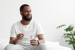 Smiling young african american man with beard hold pill and glass of water looks at empty space in bedroom interior. Covid-19 quarantine, disease treatment, health care, medicine, vitamins and therapy