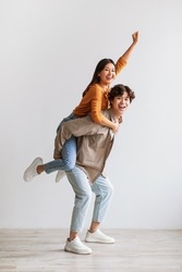 Handsome young Asian man giving piggyback ride to his girlfriend against white studio wall, full length portrait. Millennial couple having fun, spending romantic times together