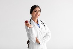 Happy pretty young indian woman doctor dentist in white coat with stethoscope hold fresh red apple on light background. Proper nutrition, vitamins, healthy diet, recommendation for perfect teeth