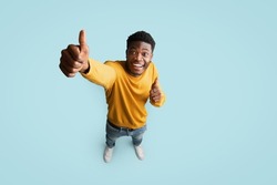 Top view of joyful handsome african american millennial guy in nice outfit showing thumb up and smiling at camera on blue studio background, recommending something exciting, copy space, full length