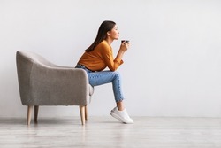 Side view of young Asian woman sitting in armchair, drinking hot aromatic coffee, relaxing against white studio wall, free space. Lovely millennial lady enjoying warm beverage on lazy day