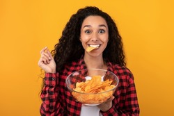 Tasty Fast Food. Portrait Of Hungry Excited Fit Female Model Eating Delicious Potato Crisps Holding Glass Bowl, Posing With Chips In Mouth Isolated Over Yellow Orange Studio Wall. Junk Meal Addiction