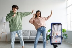 Young Asian couple recording video content, dancing on smartphone camera at home. Positive millennial boyfriend and girlfriend filming for social media, using mobile device. Vlogging concept