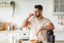 Healthy nutrition for muscle gain and weight loss concept. Athletic young Arab man drinking protein shake or milk, standing near table with healthy wholesome products at kitchen, copy space
