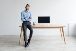 Mock Up Template. Happy Smiling Arab Man Sitting On Desk Posing With Folded Arms Showing Pc Computer With Blank Black Screen At Home Office, Free Copy Space. People, Technology, Remote Work Concept