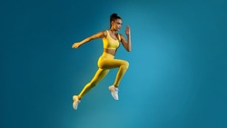 Motivated Young Lady Jumping Posing In Mid-Air Over Blue Studio Background, Looking Aside. Fitness Woman Exercising Wearing Yellow Sportswear Running In Air. Panorama, Full Length Shot