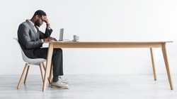 Side view of young black office employee feeling tired of working overtime, sitting at desk with laptop against white studio wall, copy space. Exhausted businessman having problem with online job