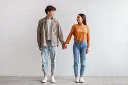 Full length portrait of millennial Asian couple smiling and looking at each other, holding hands, standing against white studio wall. Affectionate young man and woman being in love