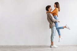 Side view of romantic young Asian guy holding and hugging his beloved girlfriend against white studio wall, copy space. Full length fo millennial spouses expressing love and affection